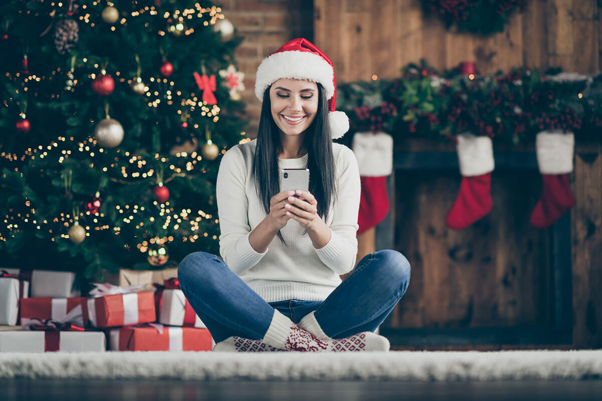 positive-girl-sir-comfort-floor-carpet-use-her-smartphone-type-text-messages-search-christmas-discounts-wear-santa-claus-hat-in-house-with-x-mas-decoration-indoors